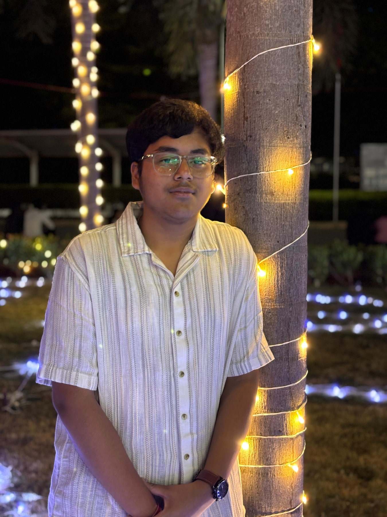 Harshit Murarka in glasses and a striped shirt, standing beside a tree trunk wrapped in warm fairy lights at night